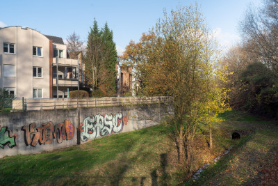 Mauer mit großflächigem Graffiti englang eines Baches, Häuser hinter der Mauer